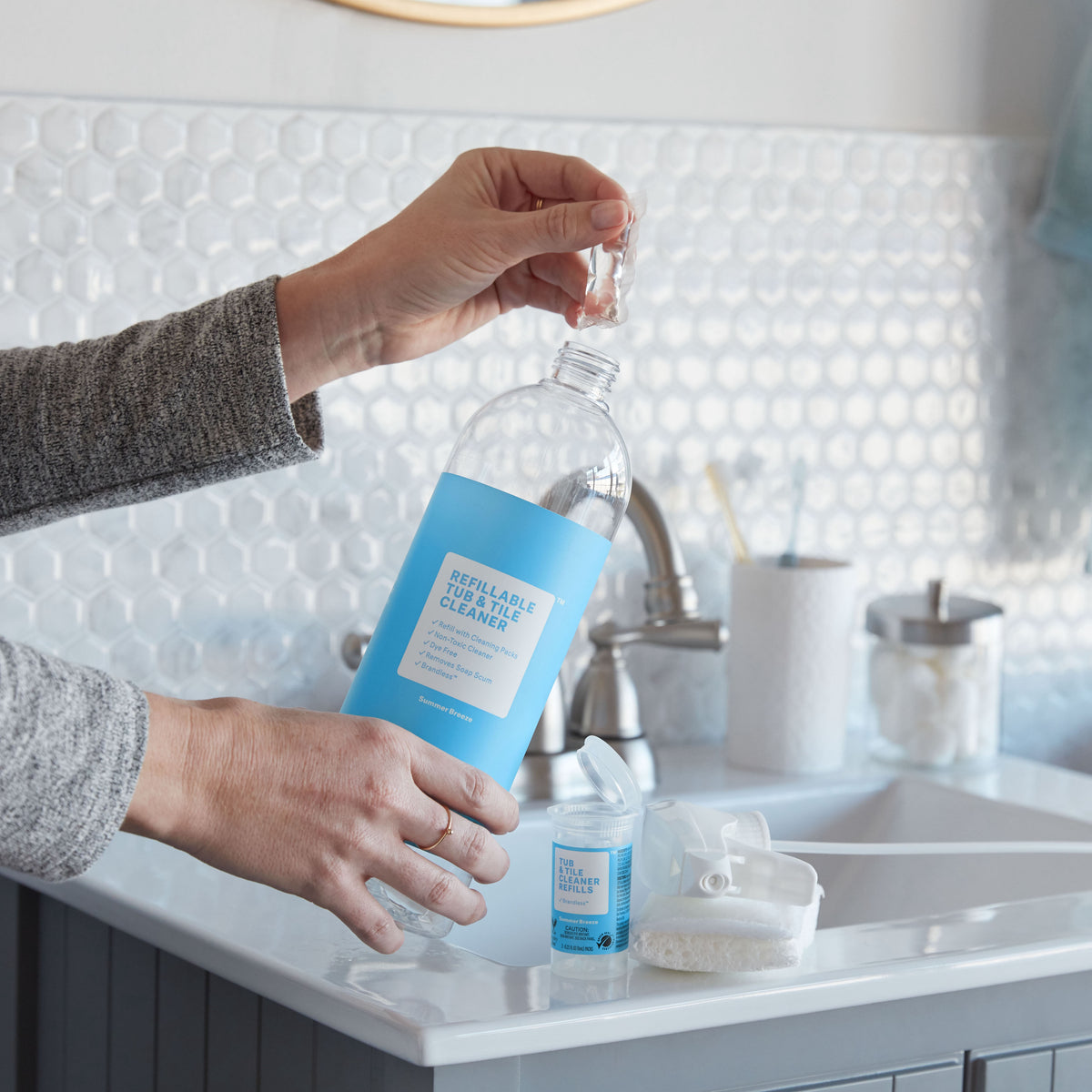 Lifestyle, hand holding an open refill container next to a spray bottle and a sink, preparing to put one packet in the spray bottle and fill with water to create one 32 oz batch of tub and tile cleaner.